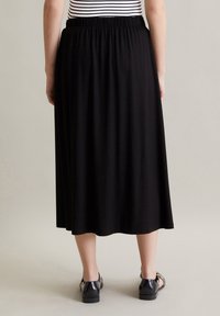 Sort, knælangt nederdel med elastisk talje. Glat stof, der svaler let fra taljen. Bæres med sorte, remmede sko.