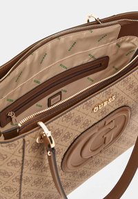 Brun håndtaske med logo mønster, glat tekstureret yderside, guldfarvede detaljer, foret inderside med grøn branding og flere lommer.