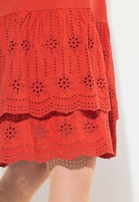 Rotes Baumwollkleid mit geschichteter Spitzenware, die florale Muster und gewellte Säume aufweist, strukturiert mit Ausschnitten und gestickten Details.