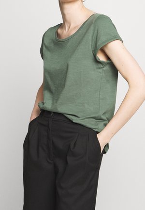 Person iført en grøn kortærmet skjorte og højtaljede sorte bukser med hænderne i lommerne, mod en ensfarvet baggrund.