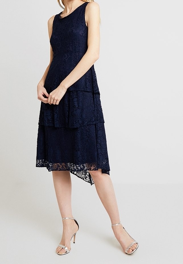 Femme portant une robe mi-longue marine en dentelle sans manches avec jupe à volants et escarpins argentés à bride à la cheville, debout devant un fond uni.