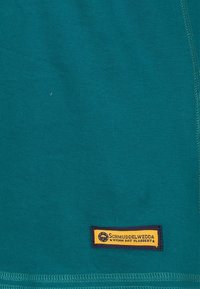 Tyrkysová látka s žlutou obdélníkovou etiketou blízko dolního okraje, na které je černý text a malé kulaté logo na levé straně.