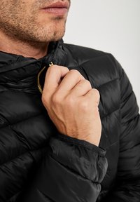 En närbild av en man i en svart pufferjacka som justerar dragkedjan med en hand, vilket avslöjar jackans eleganta design och krage.