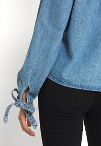 Camisa de denim azul claro con las mangas atadas con un lazo por encima de la muñeca, sobre pantalones negros ajustados, en una persona de pie.