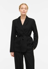 Blazer nero in tessuto morbido, con scollo a V, modello incrociato, chiusura laterale a laccio e due tasche frontali, abbinato a pantaloni coordinati.