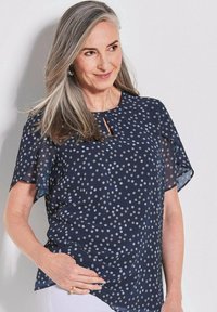 Marineblauwe blouse met korte, doorzichtige mouwen, voorzien van een stippenpatroon in lichtgrijs. Opvallend sleutelgatdetail bij de halslijn. Soepele textuur.