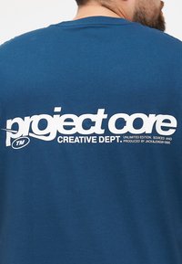 Sininen puuvillat-paita, jossa on suuri valkoinen graafinen printti, jossa lukee "project core CREATIVE DEPT." ja bränditiedot takana.