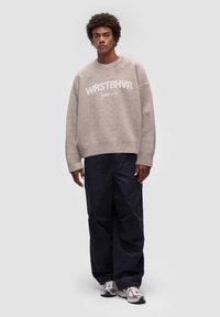 Beżowy sweter dziany z napisem "WRSTBVR" i "BERLIN" w białym kolorze, w połączeniu z luźnymi czarnymi spodniami cargo i szarymi sneakersami. Gładka struktura i luźny krój.
