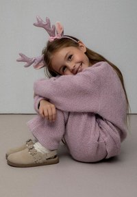 Jasnoróżowy sweter i spodnie wykonane na drutach, o przytulnej fakturze. Uzupełnione o błyszczące rogi renifera i beżowe buty wsuwane z kwiatowymi akcentami.