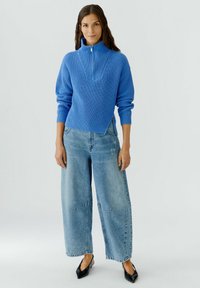Blauer Strickpullover mit hohem Kragen und Reißverschluss vorne, kombiniert mit leichten, hellblauen weit geschnittenen Jeans mit dezenten, abgenutzten Details.