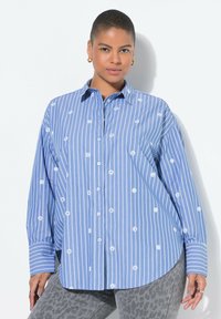 Blau-weiß gestreiftes Button-Down-Shirt mit gestickten Blumenmotiven; lange Ärmel, Kragen und abgerundeter Saum. Getragen mit grauen gemusterten Hosen.