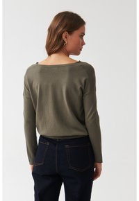 Pulover tricotat de culoare verde măsliniu, cu o croială lejeră, decolteu larg în stil barcă, umeri lăsați și mâneci lungi, împreună cu blugi de culoare albastru închis.