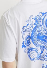 Vit bomulls t-shirt med en detaljerad blå tuppgrafik på baksidan, omgiven av intrikata blommönster. Korta ärmar, avslappnad passform.