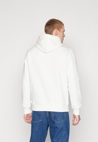 Un sweat à capuche blanc avec une capuche spacieuse, des poignets et un ourlet côtelés, associé à un jean en denim bleu, vu de dos. Texture lisse, design simple.