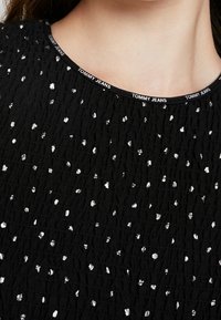 Close-up of a black textured shirt with scattered silver dots and a round neckline featuring "TOMMY JEANS" printed trim.