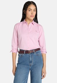 Ljusrosa skjorta med uppvikta ärmar, med en liten logotyp på bröstet. Matchas med högmidjade jeans i denim och ett brunt bälte.