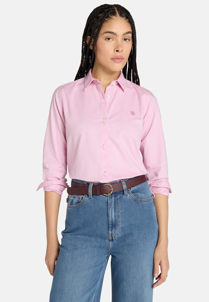 Ljusrosa skjorta med uppvikta ärmar, med en liten logotyp på bröstet. Matchas med högmidjade jeans i denim och ett brunt bälte.