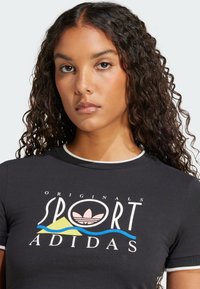 Černé přiléhavé tričko s bílým límečkem, s grafikou "ADIDAS ORIGINALS SPORT" v bílé a pastelových barvách, s křivkami v modré a žluté barvě.