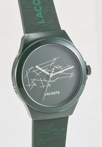 Roheline Lacoste kell, millel on ümmargune nägu, valged joonmustrid ja sobiv pehme silikoonist rihm. Näha on hõbedased käed ja logo detail.
