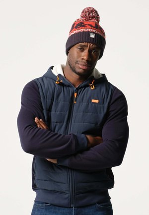 Marineblauwe gewatteerde vest met capuchon en oranje accenten, gecombineerd met een gebreide beanie met rode en grijze patronen. Model heeft zijn armen gekruist.