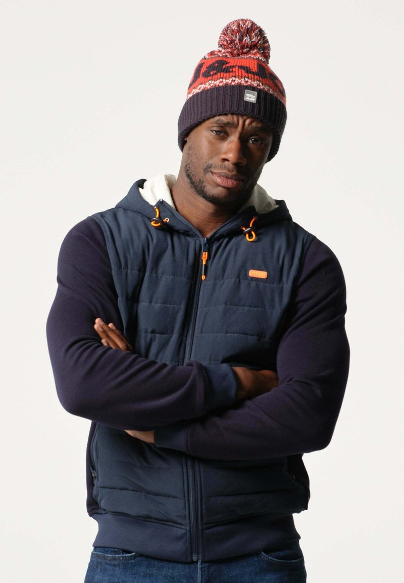 Marineblauwe gewatteerde vest met capuchon en oranje accenten, gecombineerd met een gebreide beanie met rode en grijze patronen. Model heeft zijn armen gekruist.