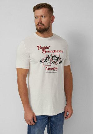 Mężczyzna z brodą, ubrany w biały T-shirt z grafiką gór i napisem "Pushin' Boundaries Country Pacemakers" oraz niebieskie dżinsy, patrzący na boki.