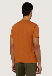 Camiseta de manga corta en algodón color naranja quemado, con cuello redondo y dobladillo recto, combinada con pantalones verde oliva, vista desde la parte de atrás.