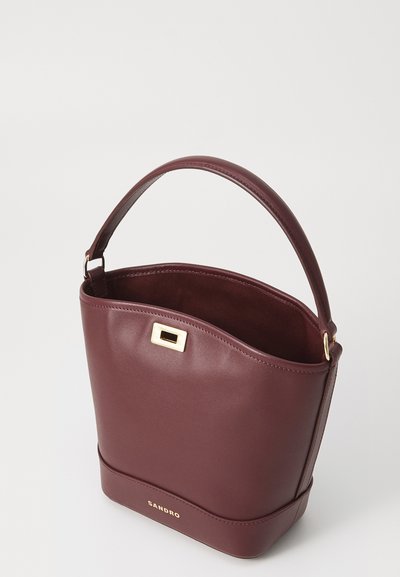 Borsa a secchiello in pelle bordeaux con una superficie liscia, forma curva, manico corto e un dettaglio in metallo dorato sulla chiusura.