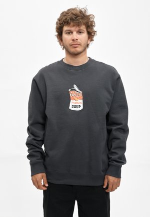 Jonge man met krullend haar draagt een donkergrijze trui met een afbeelding van een open soepblik met het etiket Hamburg Soup, staand tegen een witte achtergrond.
