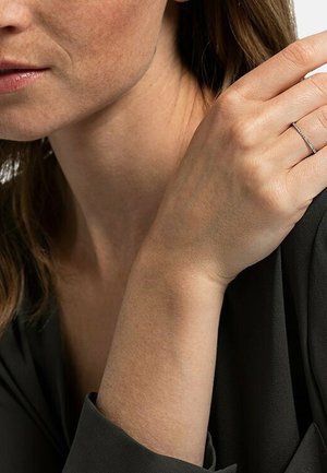 Silberner Ring mit einem zarten Design, das einen dünnen Ring und kleine Akzente aufweist. Die Hand ist anmutig vor einem dunklen Stoffhintergrund positioniert.