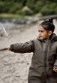 Giacca imbottita marrone con cappuccio, con polsini elasticizzati. Bambino che tiene in mano una lunga pianta secca bianca su uno sfondo naturale sfocato.