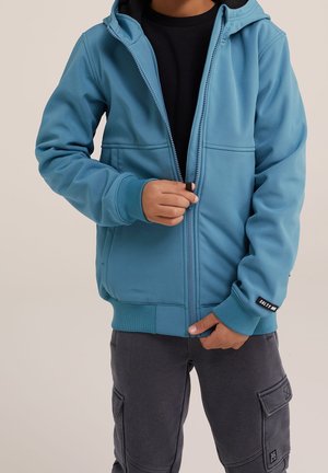 Dziecko w turkusowej kurtce z kapturem, którą odzipowuje, w połączeniu z czarną koszulką i ciemnoszarymi spodniami bojówkami.