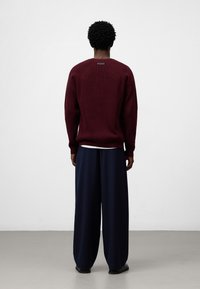 Bordeauxfarbenes Strickpullover mit geripptem Muster, hohem Kragen und Logodetail auf der Rückseite, kombiniert mit lockeren, marineblauen Hosen. Einfacher Hintergrund.