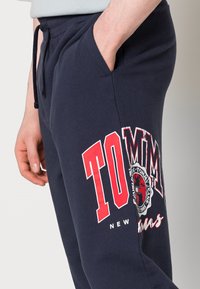 Tmavě modré tepláky s výrazným červeným motivem obsahujícím text "TOMMY" a logo. Navrženy s elastickým pasem a bočními kapsami.