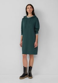 Vrouw in een donkergroene hoodie jurk over een gestreepte top en zwarte chunky laarzen, staande tegen een effen lichtgrijze muur.