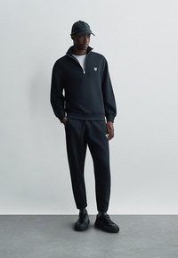 Fekete pulóver fél cipzárral és logóval, párosítva illeszkedő, beszűkített sportnadrággal és fekete sportcipővel. Finom textúrával és sportos dizájnnal rendelkezik.