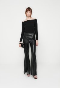 Donna che indossa un top nero scollato e pantaloni in pelle neri a vita alta e svasati, che tiene in mano una piccola borsa nera, in piedi davanti a uno sfondo bianco.