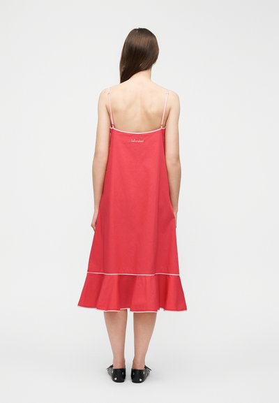 Γυναίκα που στέκεται με την πλάτη στραμμένη, φορώντας ένα αμάνικο κόκκινο μίντι φόρεμα με λεπτούς ροζ ιμάντες και μαύρα φλατ παπούτσια με τρουκς, σε απλό φόντο.