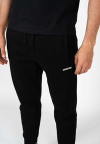 Sort sweatpants lavet af blødt stof, med en snoretræk i taljen og et lille mærke på siden. Tætsiddende omkring anklerne.