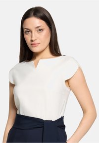 Blouse blanche à encolure en V, manches courtes, texture lisse, associée à un pantalon taille haute bleu marine avec une ceinture nouée.