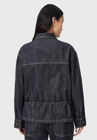 Donna con lunghi capelli scuri che indossa una giacca di denim scuro e pantaloni abbinati, vista di spalle su uno sfondo grigio uniforme.