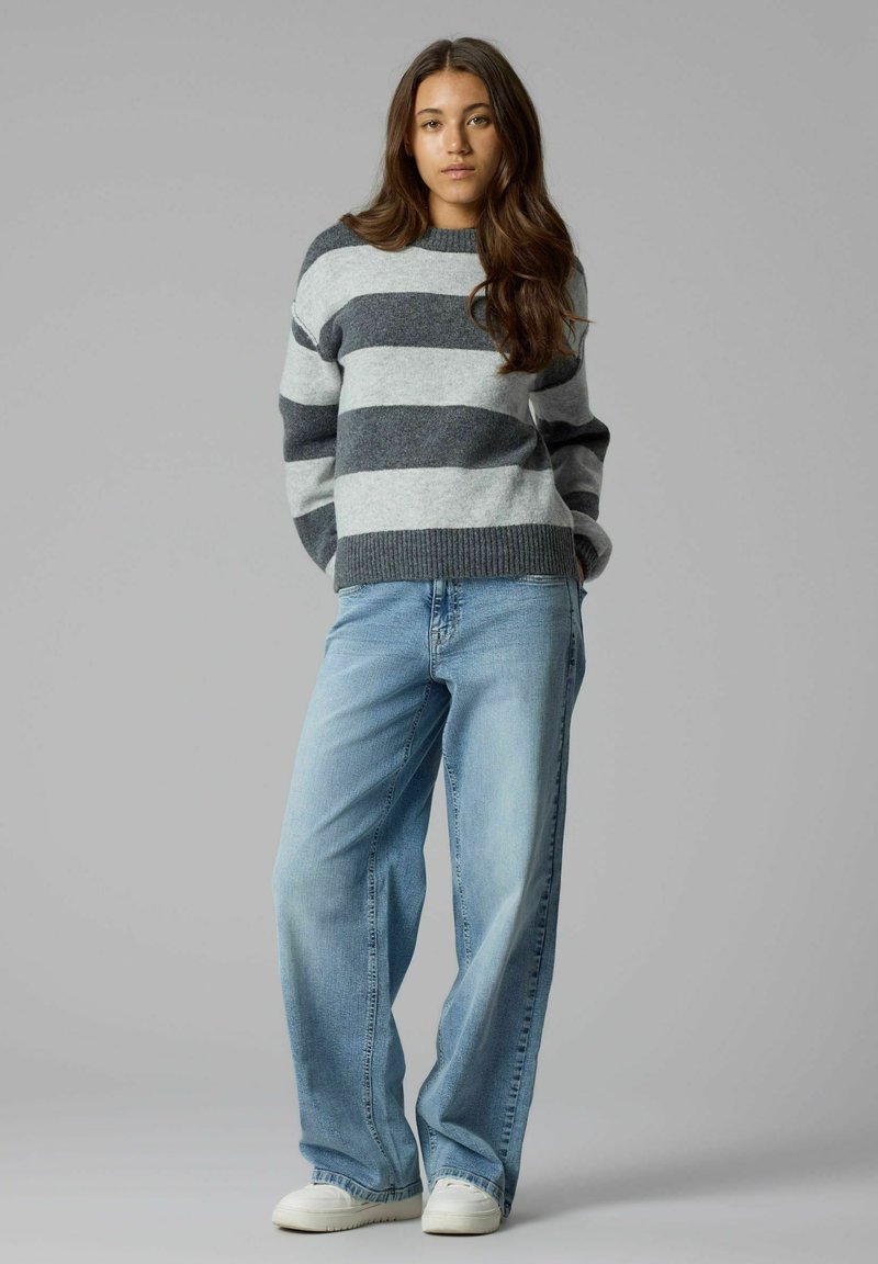 Jeune femme aux longs cheveux portant un pull rayé gris et gris foncé, un jean large bleu clair et des baskets blanches, debout devant un fond gris.