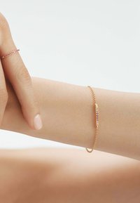 Pulsera de oro con un diseño de cadena delicada y un centro curvado que presenta pequeñas piedras preciosas rosas, llevada en un brazo desnudo.