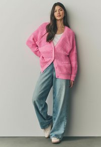 Lyserød strikket cardigan med løs pasform, strukturerede mønstre og V-hals. Sættes sammen med brede blå jeans og hvide sneakers.