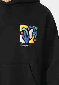 Sudadera negra con un bolsillo frontal, que presenta un diseño gráfico abstracto multicolor en la parte izquierda del pecho. El material parece suave.