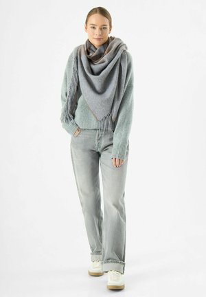 Grijze en bruine oversized sjaal met franjes, gedragen over een lichtblauw sweater. Gecombineerd met lichtgrijze jeans en witte sneakers.