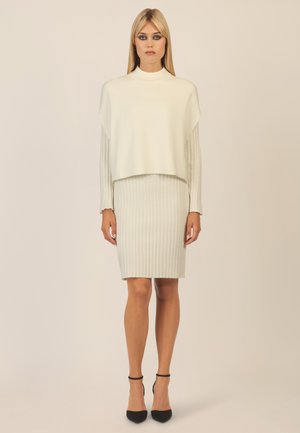 Hellgrauer Strickpullover mit hohem Kragen und verkürztem Design, kombiniert mit einem passenden, gerippten Bleistiftrock. Schwarze Riemchensandalen mit Absatz runden den Look ab.