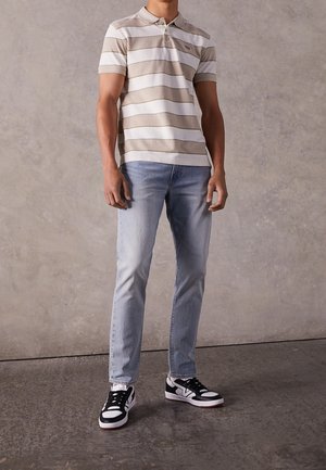 Man draagt beige-wit gestreept poloshirt, lichtblauwe spijkerbroek en zwart-witte sneakers, staand tegen een grijze, gestructureerde muur.