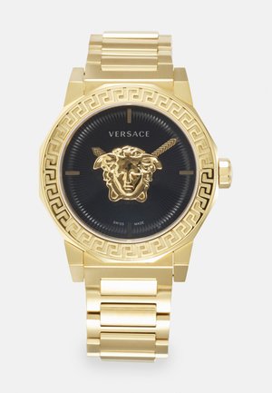 Montre octogonale dorée avec un cadran noir, arborant un emblème de Méduse et un motif grec complexe sur la lunette. Fabriquée en Suisse.