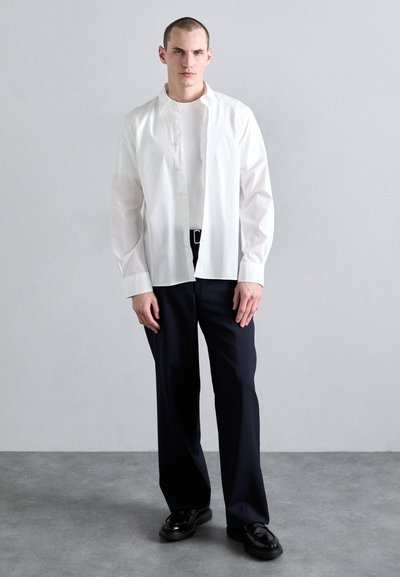 Chemise blanche à boutons avec des manches longues, superposée à un t-shirt blanc ajusté. Associée à un pantalon large bleu marine foncé et des chaussures noires.